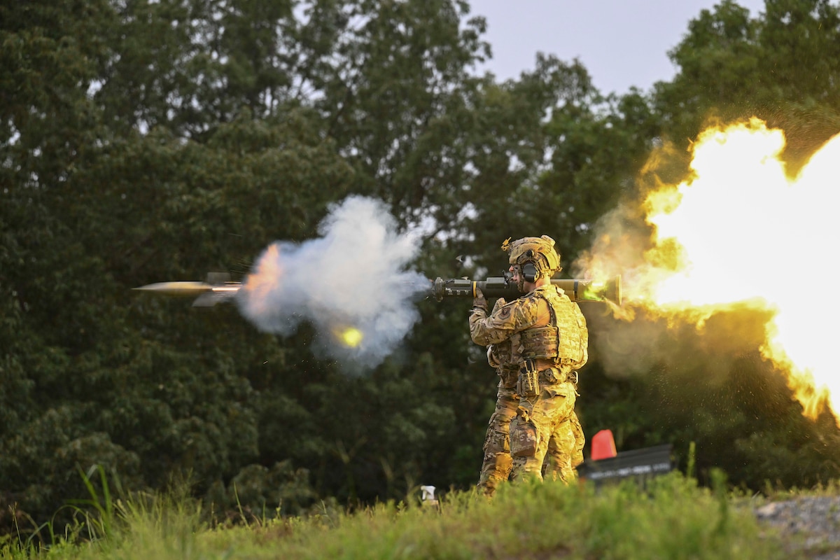 Two service members wearing camouflage stand close to each other as one fires a shoulder-launched missile that creates smoke in front and fire behind the launcher.