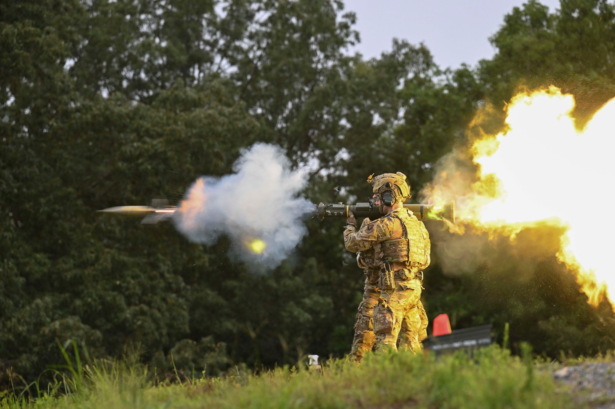 Two service members wearing camouflage stand close to each other as one fires a shoulder-launched missile that creates smoke in front and fire behind the launcher.