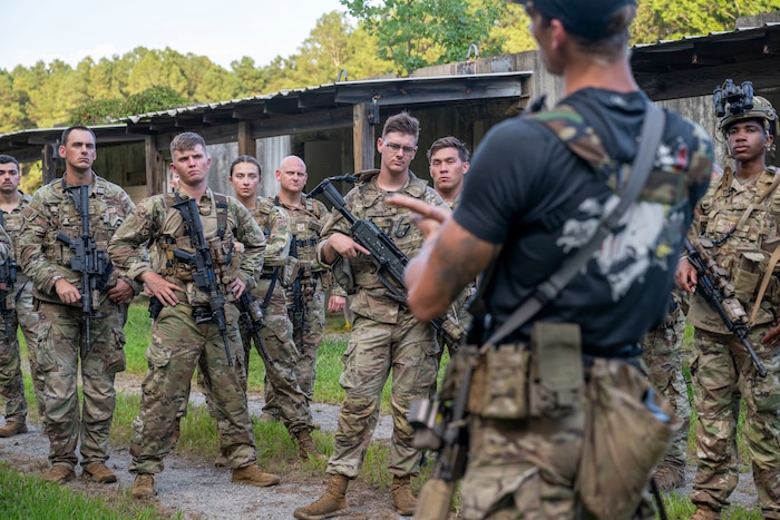 Several people wearing camouflage uniforms and holding rifles stand together and listen to a person in a black T-shirt and camouflage pants speak in the foreground.