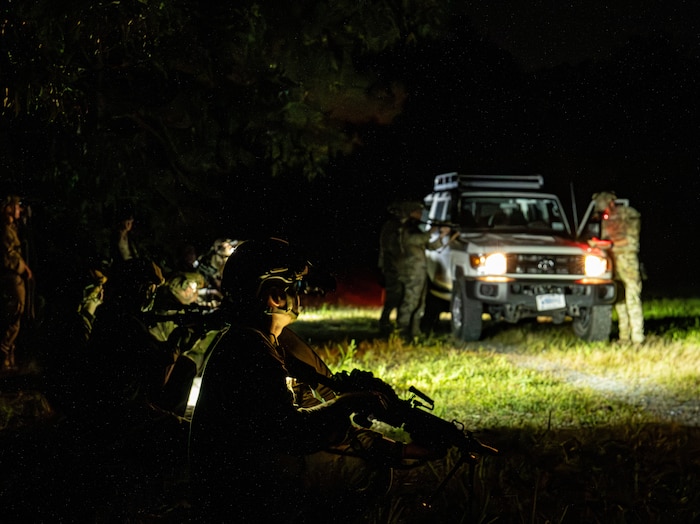 Two soldiers in camouflage uniforms stand beside a vehicle at night. The vehicle has its headlights on, and there are additional soldiers in similar attire kneeling in the foreground. The area in the foreground is dark, and the soldiers are holding rifles.