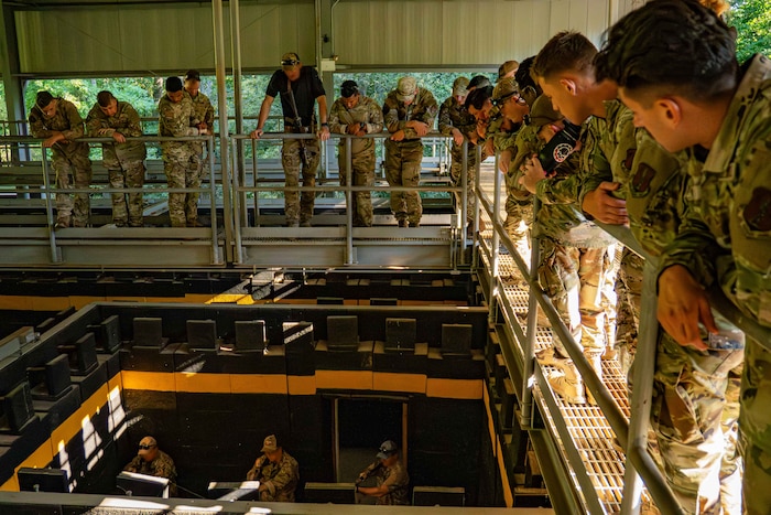 Service members in camouflage uniforms line a catwalk and look down at other service members in similar attire as they walk through a room with an open roof. The people on the ground are holding rifles as they walk.