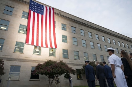 A large American flag hangs off the side of a large, five-story building at sunrise. Service members in military attire face the flag and stand at attention.