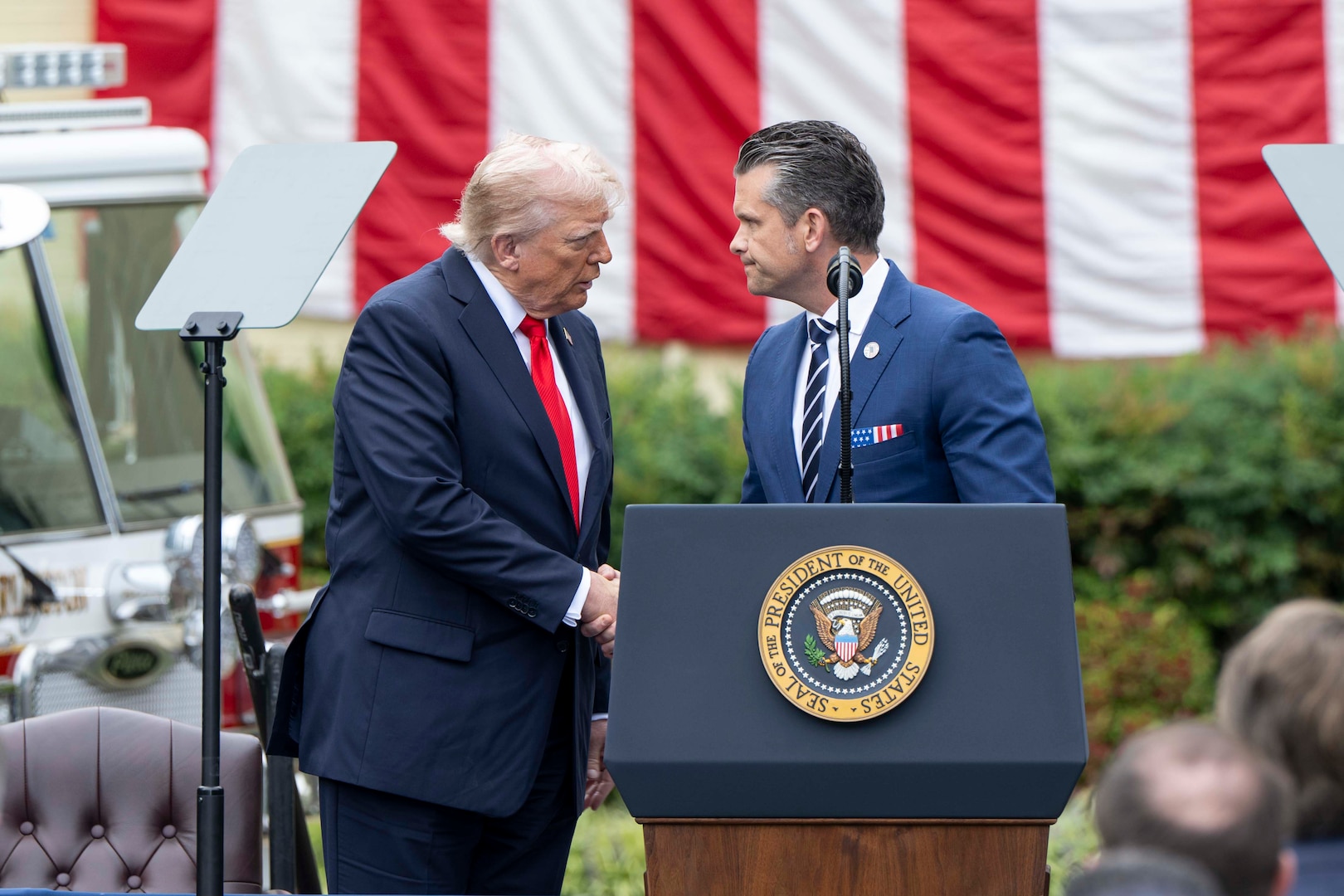 The secretary of war and the president shake hands on a stage.