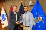Two men shake hands in front of flags.