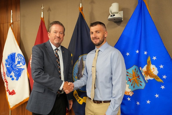 Two men shake hands in front of flags.
