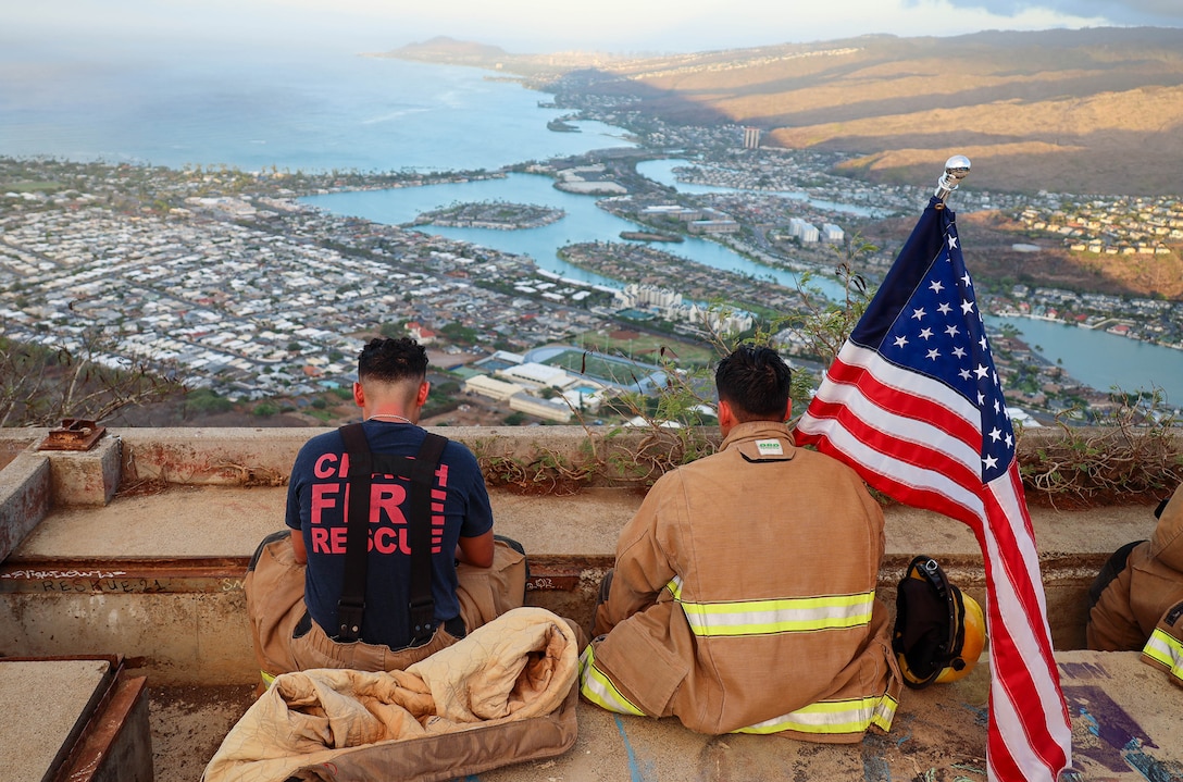 Two Marines seen from behind, one in firefighting gear holding a U.S. flag, sit on a platform overlooking coastline below.