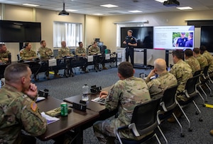 A uniformed police officer stands at the front of a classroom with a TV screen on. About a dozen people wearing military camouflage uniforms are seated at tables lining the outside of the room.