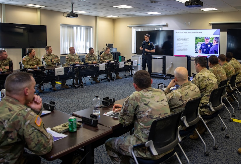 A uniformed police officer stands at the front of a classroom with a TV screen on. About a dozen people wearing military camouflage uniforms are seated at tables lining the outside of the room.