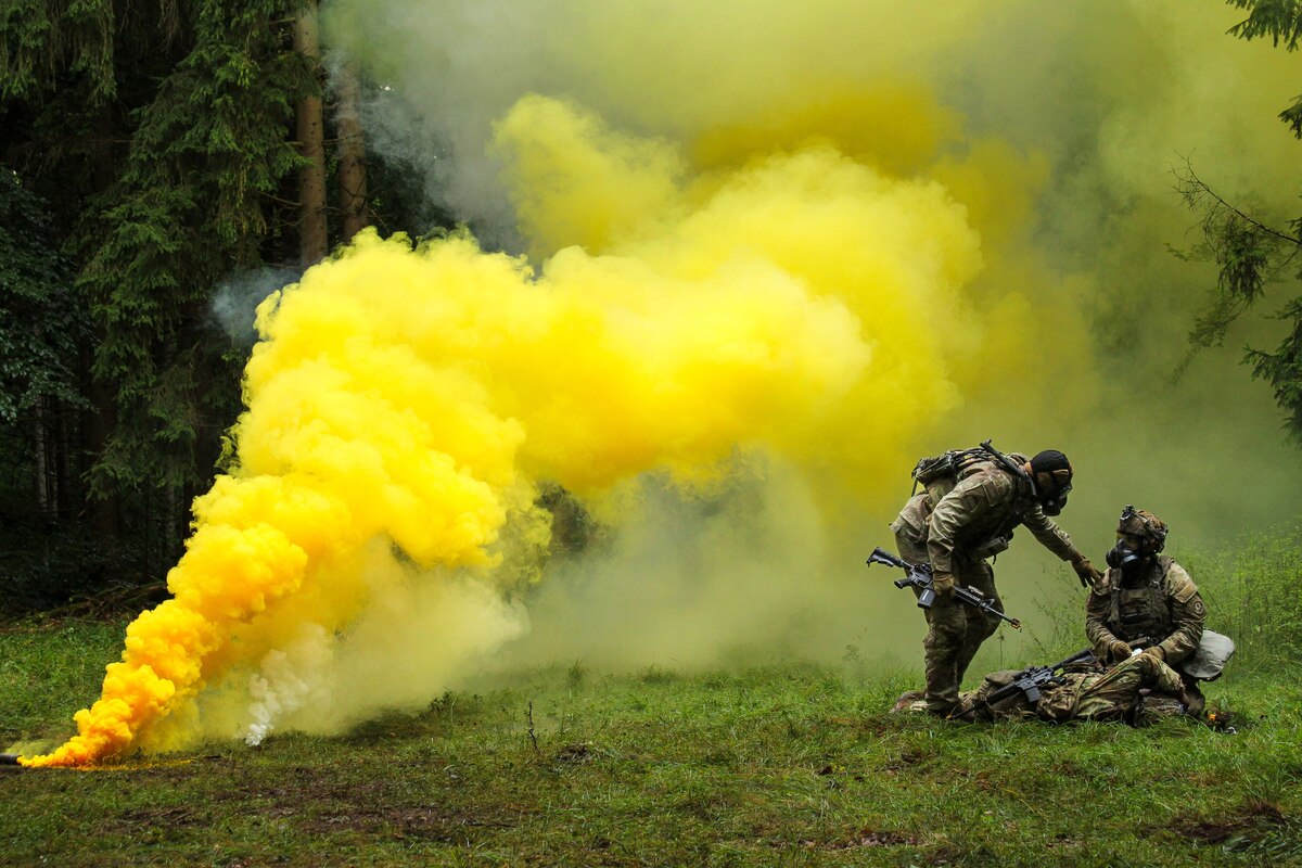 Two soldiers wearing camouflage military uniforms and gas masks provide medical assistance to another soldier in similar attire lying on the ground during an exercise. Yellow smoke billows in the background to simulate a chemical attack.