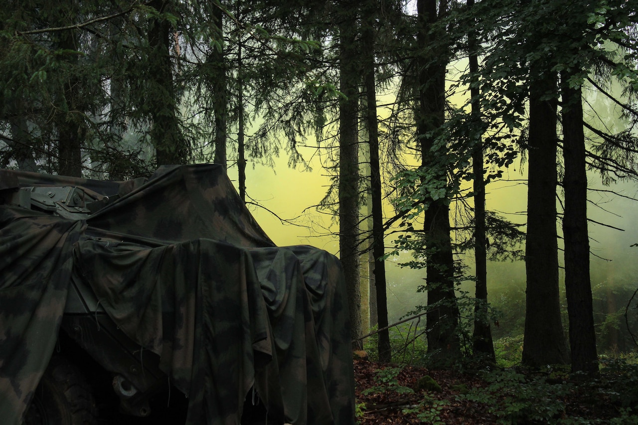 A military tent in a dark wooded area with yellow smoke in the background is shown.