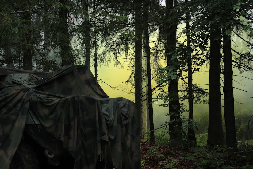 A military tent in a dark wooded area with yellow smoke in the background is shown.