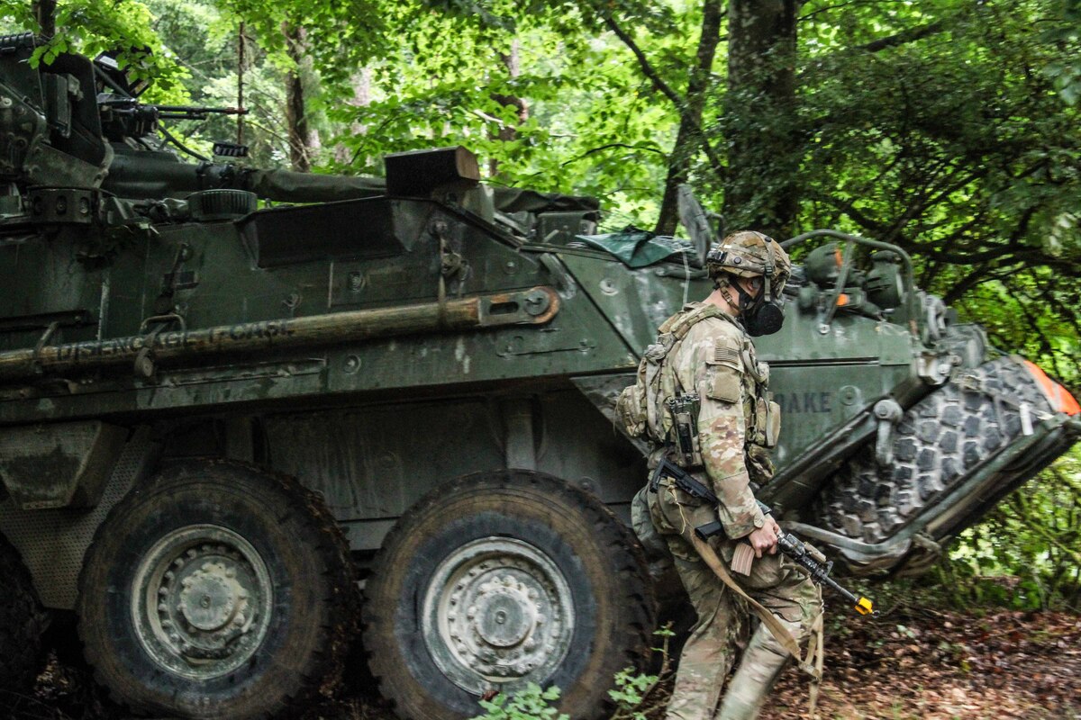 A soldier wearing a camouflage military uniform and gas mask walks in front of a military tank.