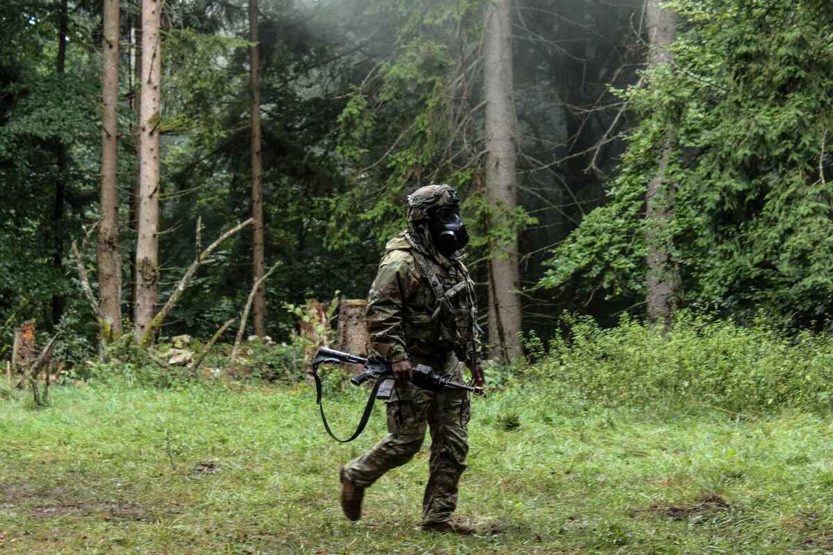 A soldier wearing a camouflage military uniform and gas mask walks in a wooded area. The soldier is carrying a rifle in their right hand.