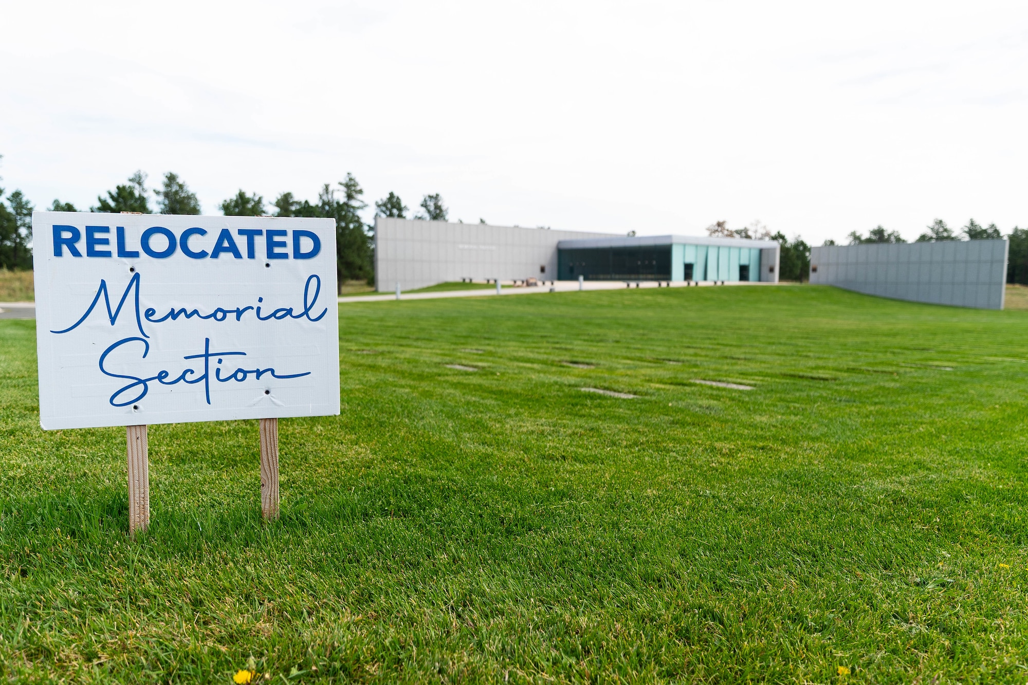 A photo of a sign that reads " Relocated memorial section". The sign is staked in the ground outside of the cemetery  building.
