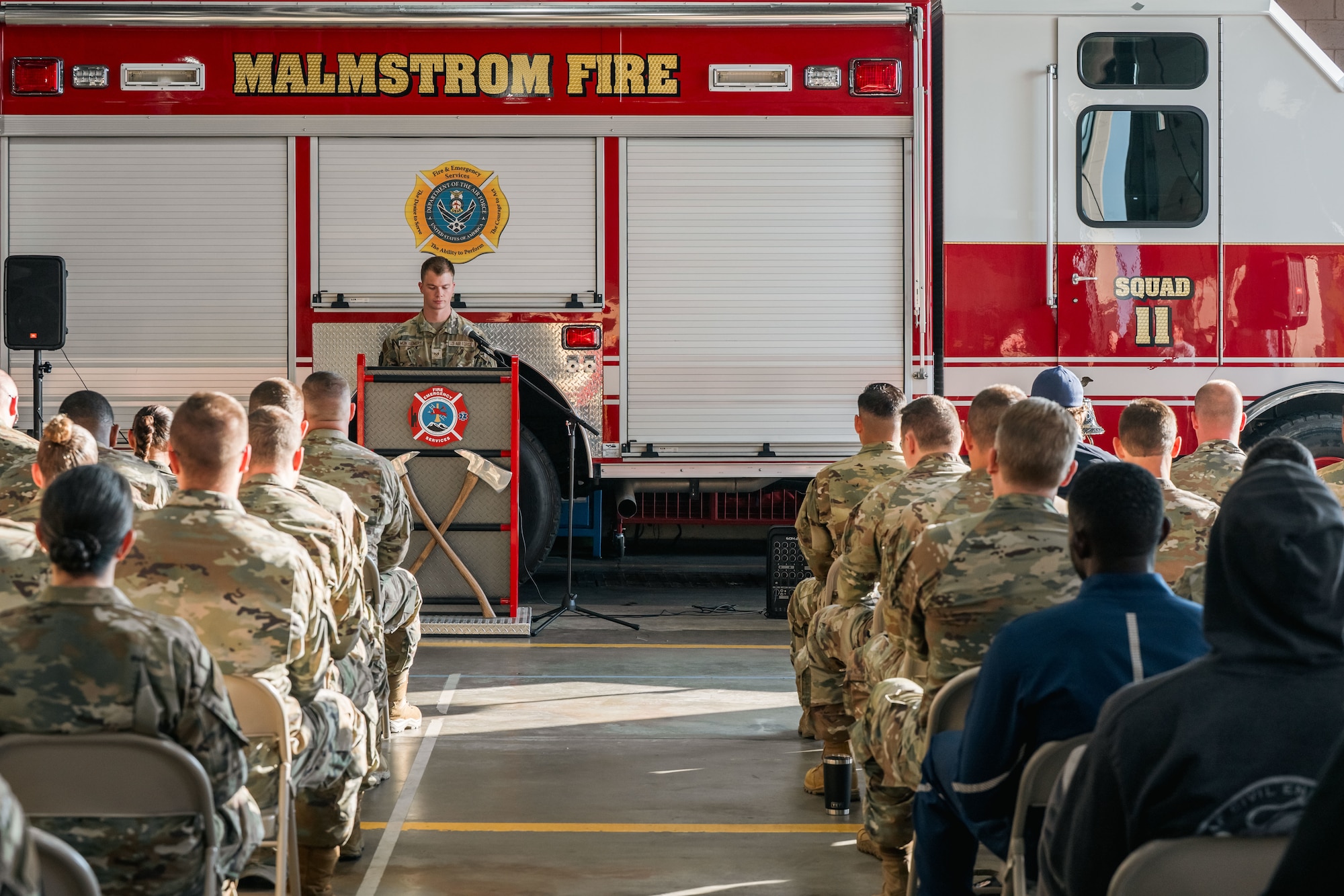 a wide shot of a group of people sitting and listening to a speaker with a firetruck in the background.