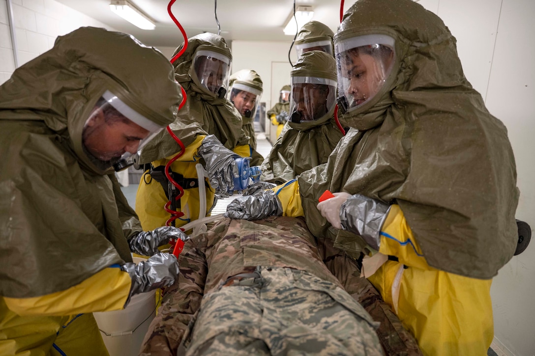 Airmen wearing hazmat suits cut clothing from a training mannequin in a bright, white room.