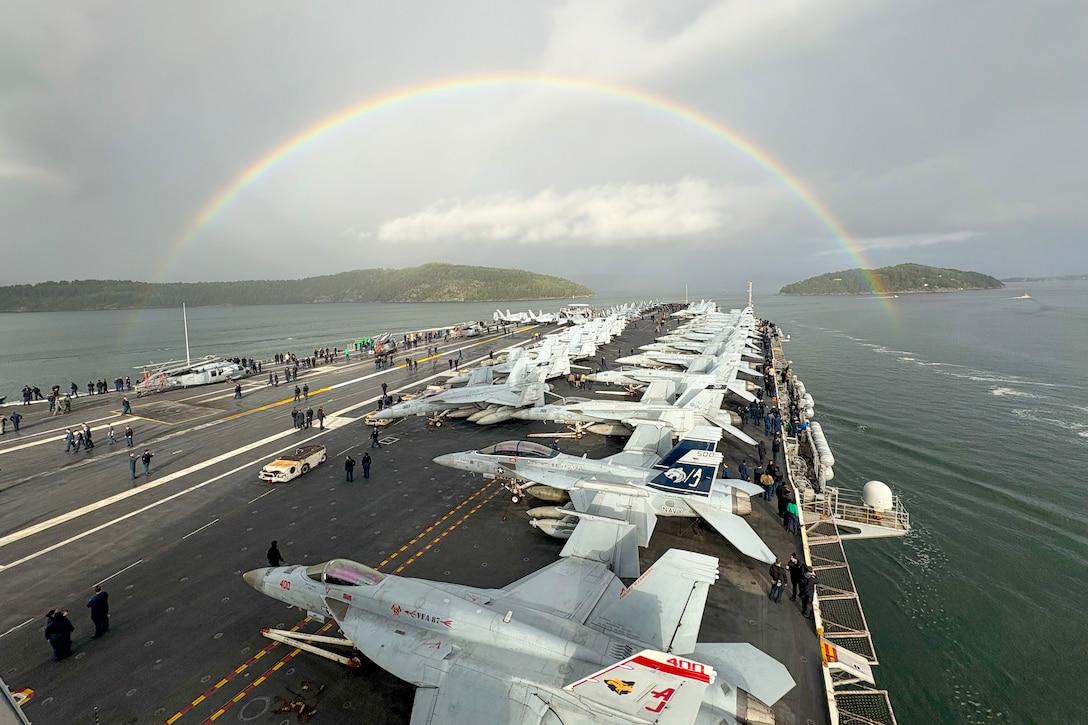 Sailors work on the flight deck of a ship near aircraft as it transits a body of water under a rainbow.