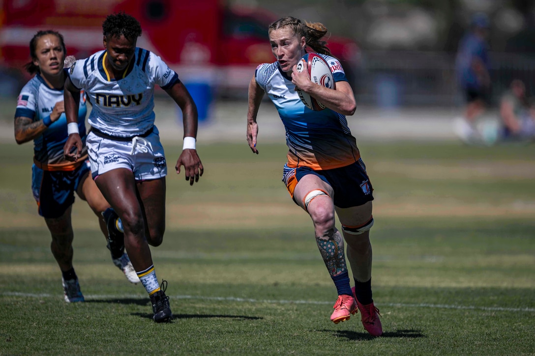 A Coast Guard rugby player runs with the ball as opposing team members run behind them on a field against blurred background.