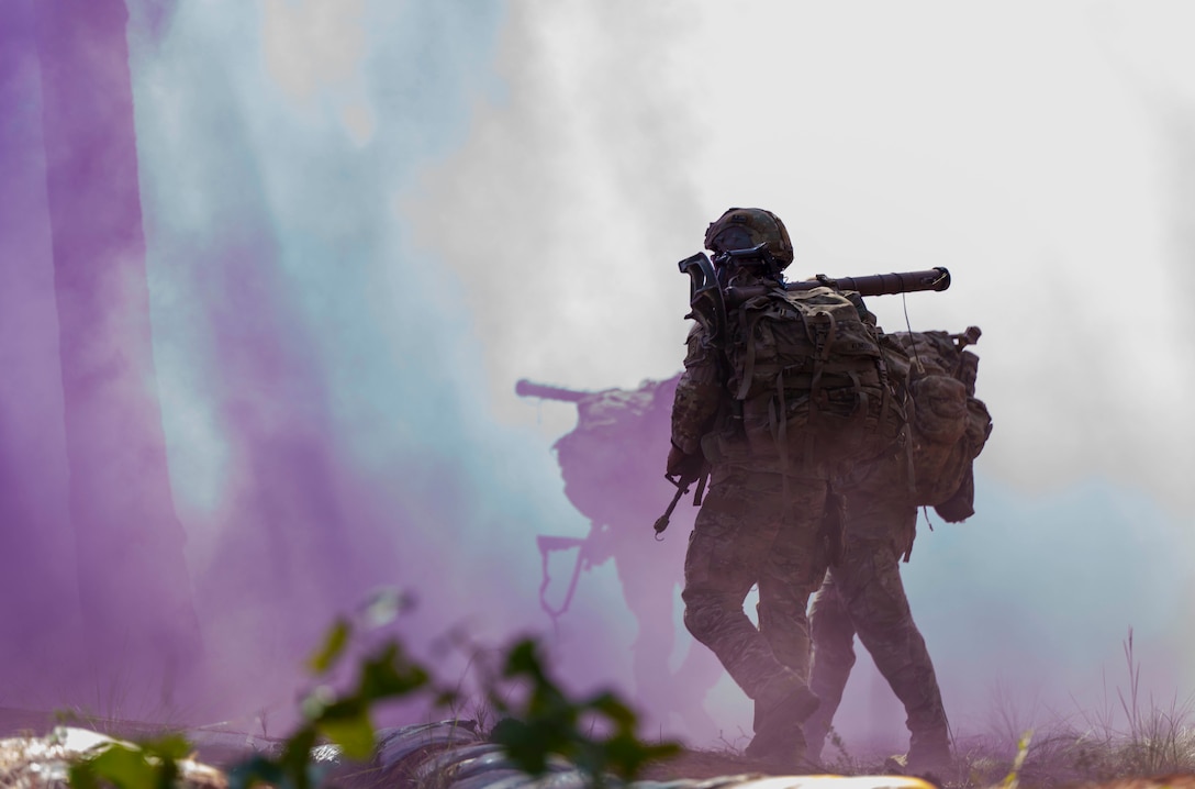 Three soldiers in full tactical gear and backpacks run through purple smoke outside during the day.
