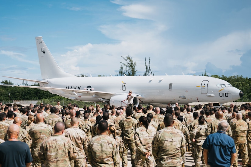 A large group of people wearing camouflage military uniforms watches a man in casual attire give a speech while standing on a platform. There is a large miliary aircraft in the background.