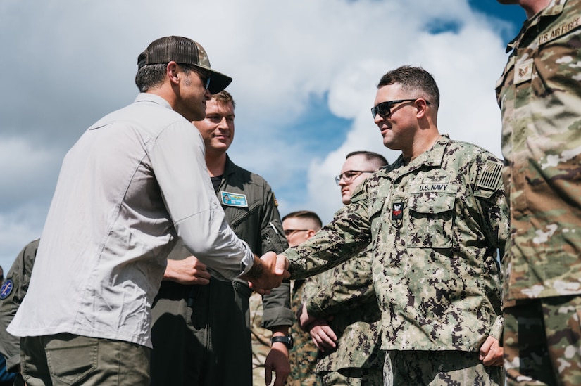 A man wearing casual attire and a hat shakes hands with a man wearing a military camouflage uniform and sunglasses. There are other service members wearing military camouflage uniforms standing next to the men shaking hands.