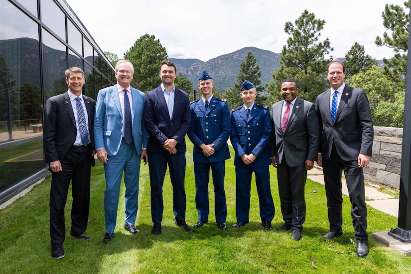 Five individuals wearing business attire and two wearing military dress uniforms stand shoulder to shoulder outdoors.  In the background are evergreen trees and mountains.