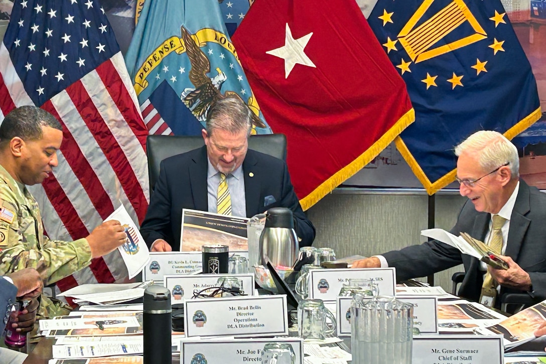 Two men wearing suits and ties and a man wearing a camouflage military uniform sit at a large conference table with military service flags behind them.