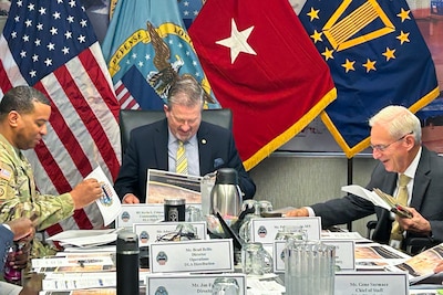 Two men wearing suits and ties and a man wearing a camouflage military uniform sit at a large conference table with military service flags behind them.