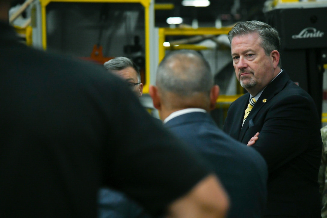 A man wearing a suit and tie stands with his arms crossed looking toward a group of people.