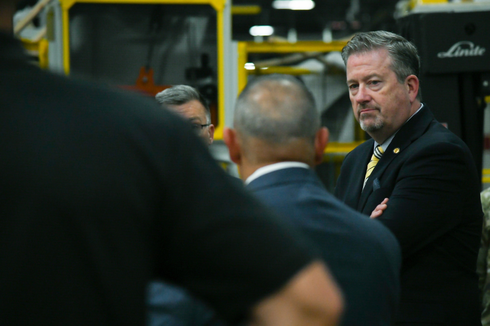 A man wearing a suit and tie stands with his arms crossed looking toward a group of people.