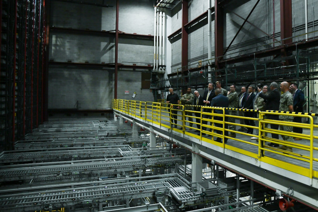 A large group of people stand behind a yellow protective railing looking a high-rise storage system inside of a large warehouse.