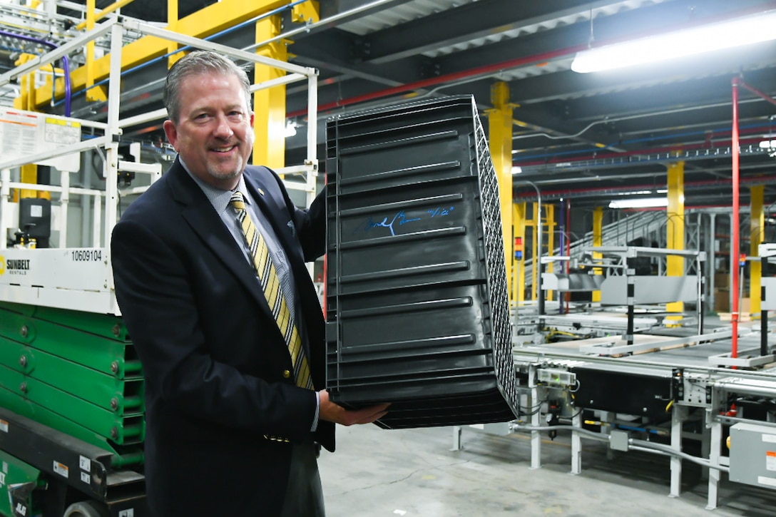 A man wearing a suit and tie holds up a signed large, black, plastic tote while standing inside of a large warehouse.