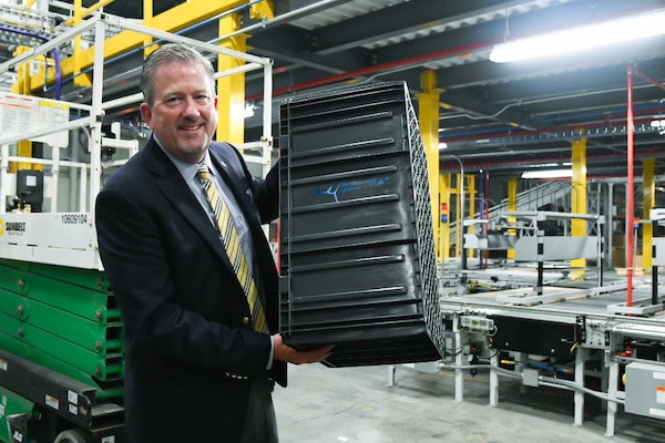A man wearing a suit and tie holds up a signed large, black, plastic tote while standing inside of a large warehouse.
