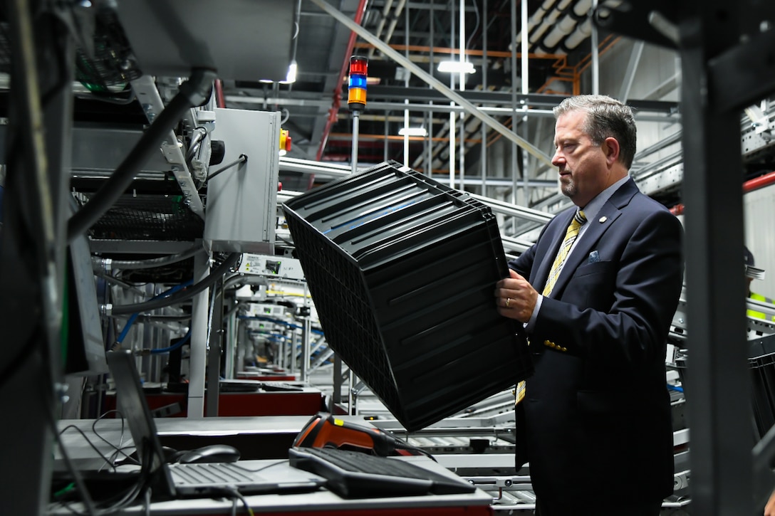 A man wearing a suit and tie loads a plastic tote into an automated storage and retrieval system inside of a large warehouse.