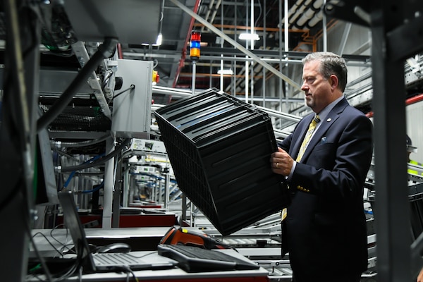 A man wearing a suit and tie loads a plastic tote into an automated storage and retrieval system inside of a large warehouse.