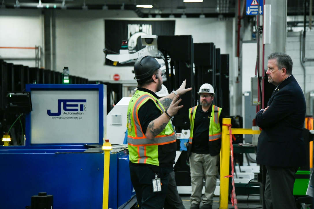A man wearing a hardhat and reflective safety vest gestures with his hands while speaking to a man wearing a suit and tie while standing inside a large warehouse.