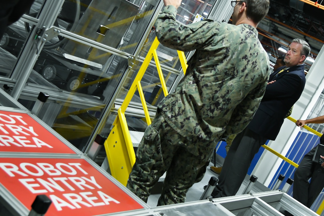 A man wearing a camouflage military uniform gestures with his hand wile speaking to a man in a suit and tie while standing in a large warehouse. There is a red sign to the left of them with white lettering that reads "robot entry area."