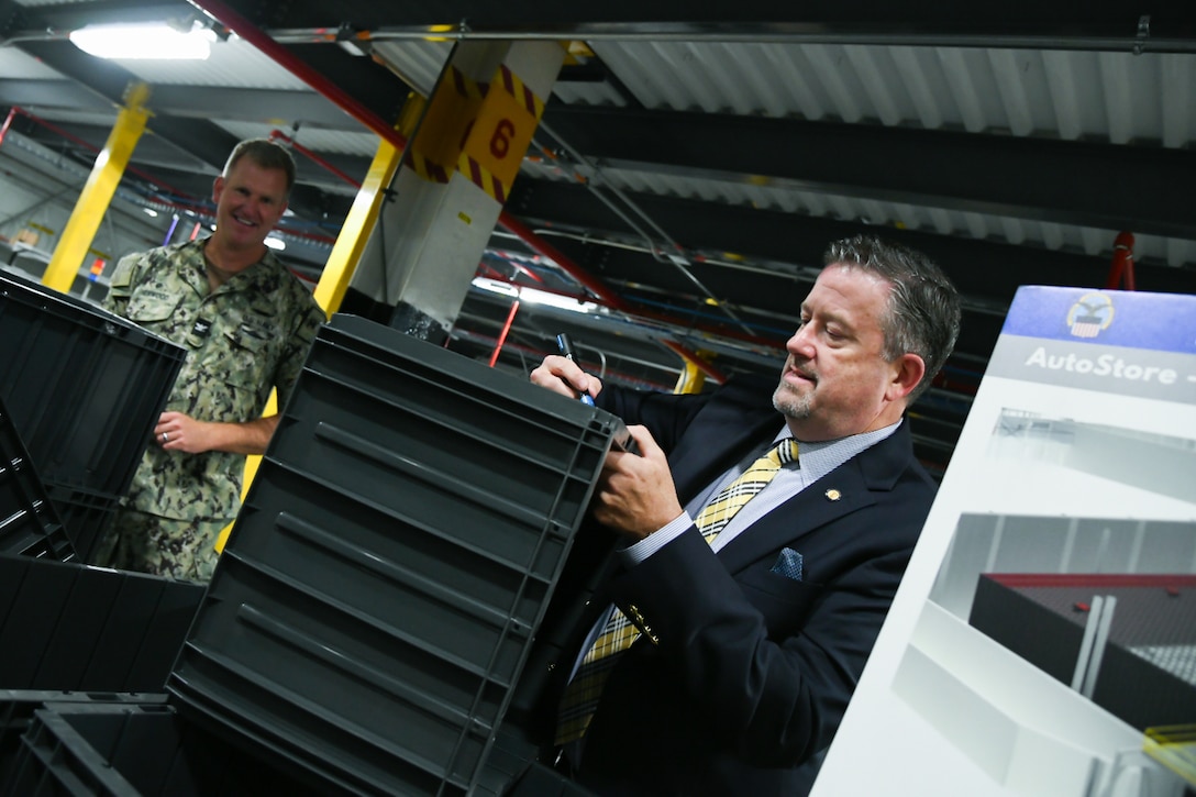 A man wearing a suit and tie signs a plastic tote with a marker standing inside of a warehouse.
