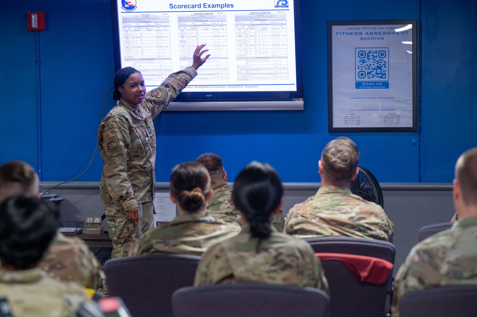 U.S. Air Force Tech. Sgt. Imani Williams, 375th Force Support Squadron fitness and sports operations manager, points to a large screen displaying a physical fitness assessment score chart when instructing a class of augmentees at Scott AFB.