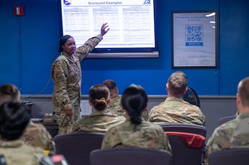 U.S. Air Force Tech. Sgt. Imani Williams, 375th Force Support Squadron fitness and sports operations manager, points to a large screen displaying a physical fitness assessment score chart when instructing a class of augmentees at Scott AFB.