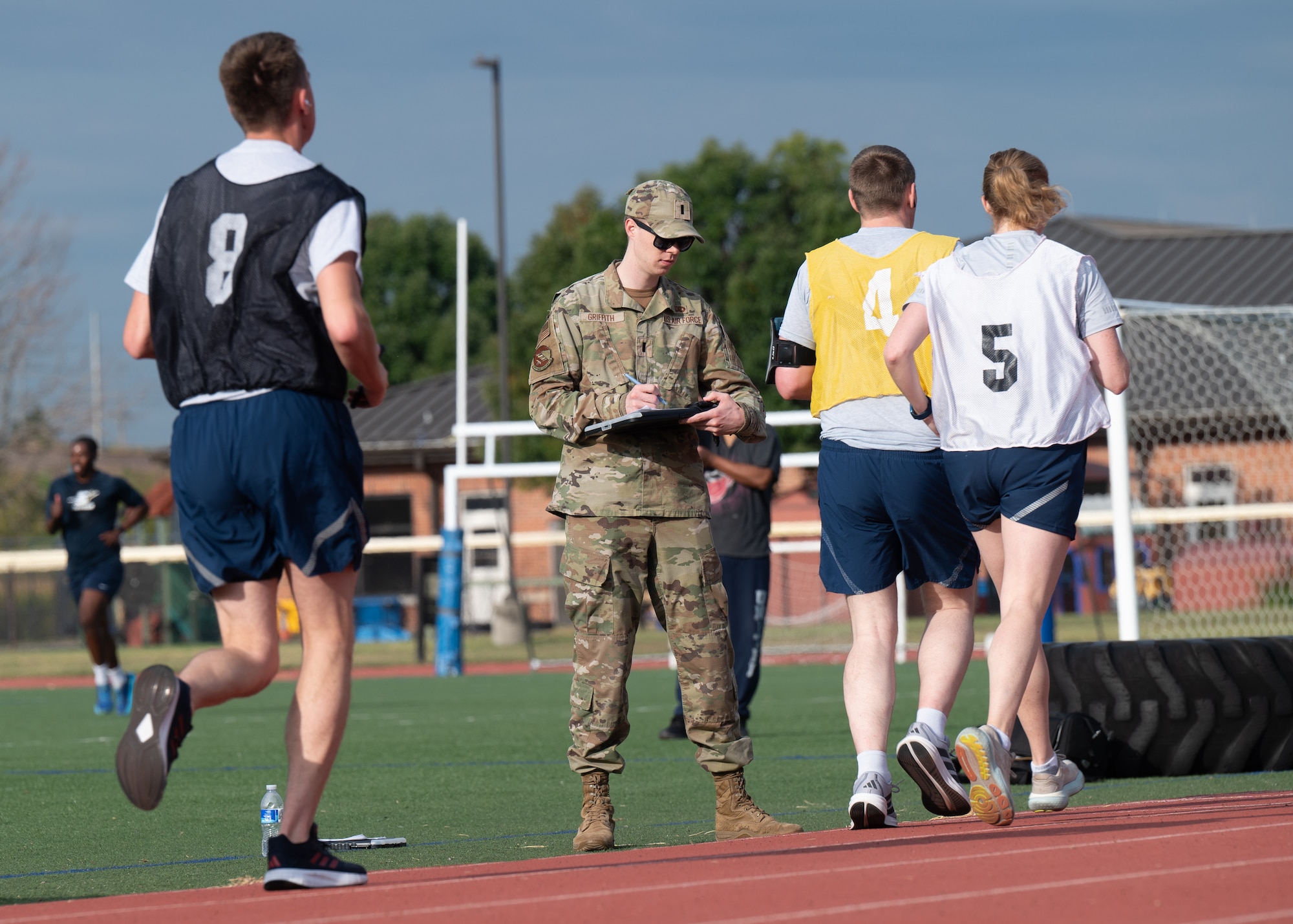 Griffith stands on the grass inside a track while writing on a clipboard. In the foreground, three Airmen wearing numbered jerseys—No. 4 in yellow, No. 5 in white, and No. 8 in black—run laps around the track.