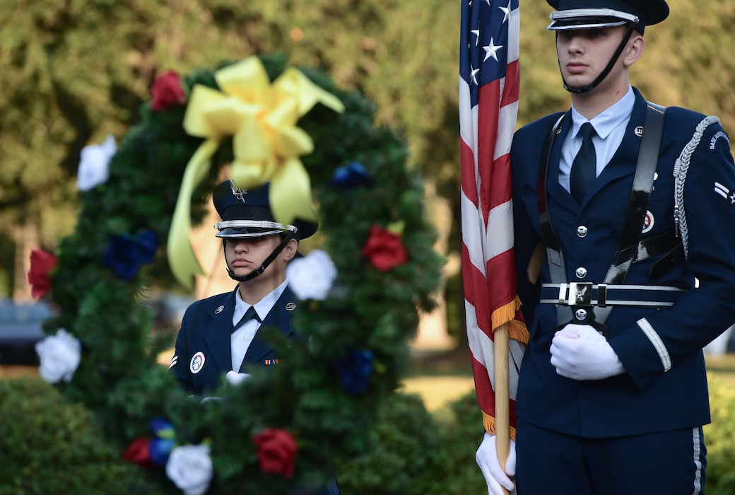 Two people wearing military uniforms stand behind a memorial wreath.