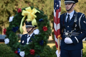 Two people wearing military uniforms stand behind a memorial wreath.