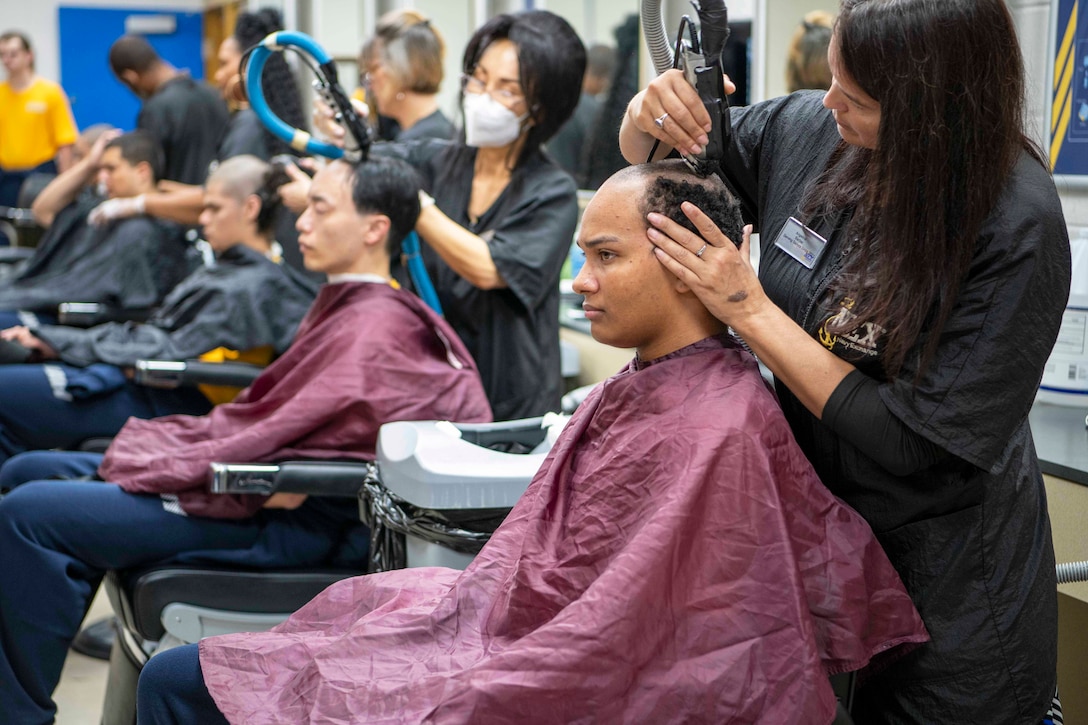Seated Navy recruits wear barber capes while getting a haircut in a salon.