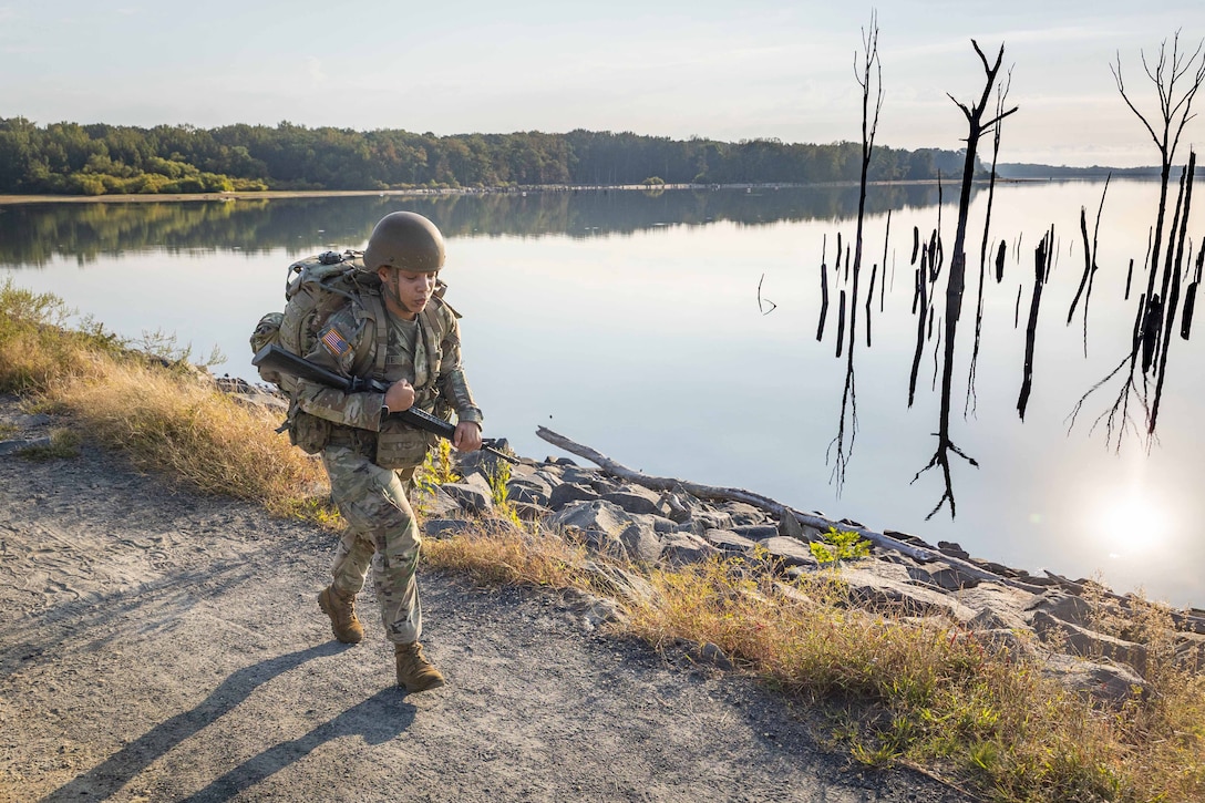 A soldier in tactical gear rucks through a wooded area near a body of water.