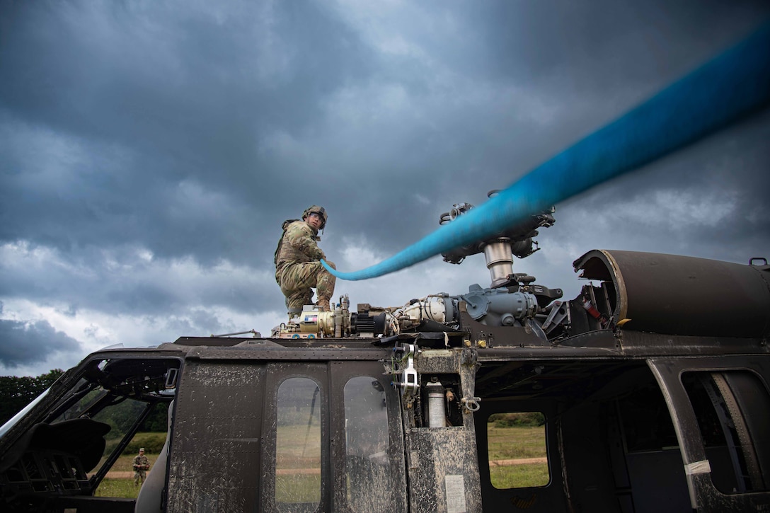 A soldier kneels on a helicopter while holding a blue line under a gray sky.