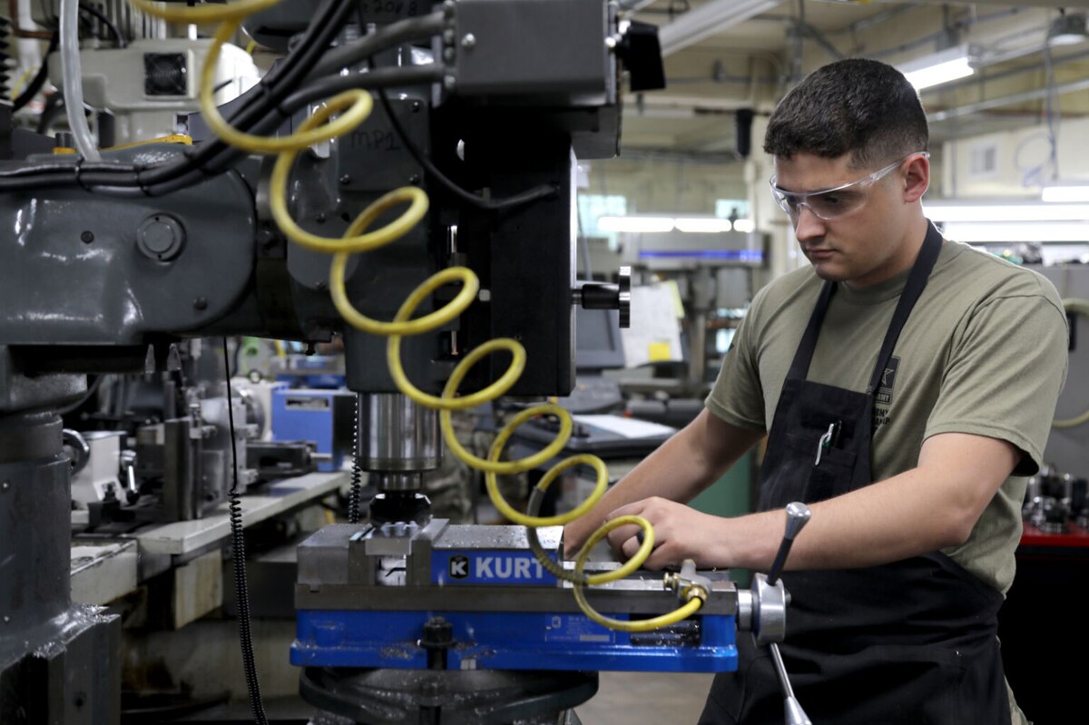 A man wearing eye protection, a green T-shirt and black apron, works on a weapon with a large piece of equipment.