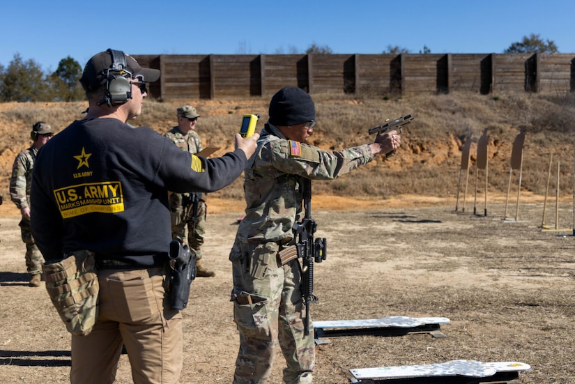 A man wearing a black sweatshirt, khaki slacks, hearing protection and a baseball cap, holds up a measuring device as a man dressed in a military camouflage uniform aims a weapon at a target not seen in the image. There are two men dressed in military camouflage uniforms standing in the background.