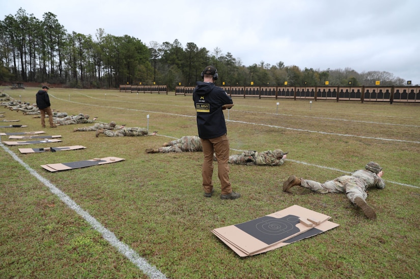 Two people dressed in black tops and khaki slacks and wearing hearing protection, stand and watch as people dressed in military camouflage uniforms lay on the ground in the prone position aiming weapons at targets in the background.