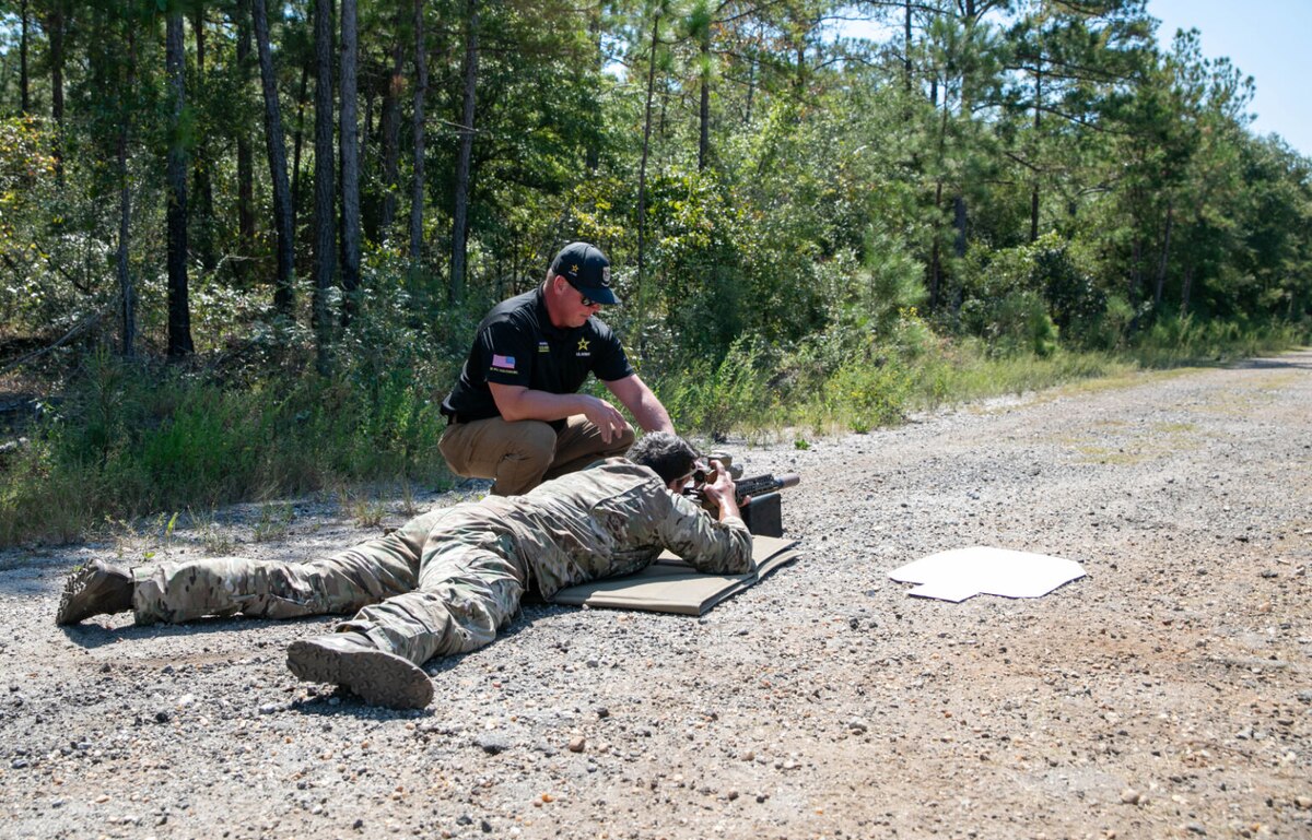 A person dressed in a military camouflage uniform lays in the prone position aiming a weapon to a target outside of the image. A man dressed in a black T-shirt and khaki slacks is kneeling next to the person in uniform.